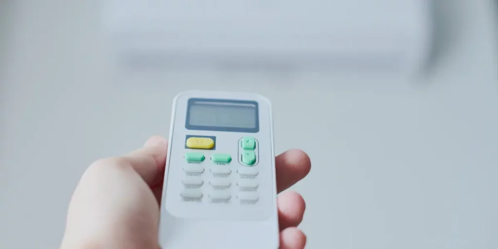 A person holding a white remote control with a blue backlight, pointing it towards a Tosot air conditioner mounted on a white wall. The air conditioner has a horizontal slatted vent and a small digital display on the right side